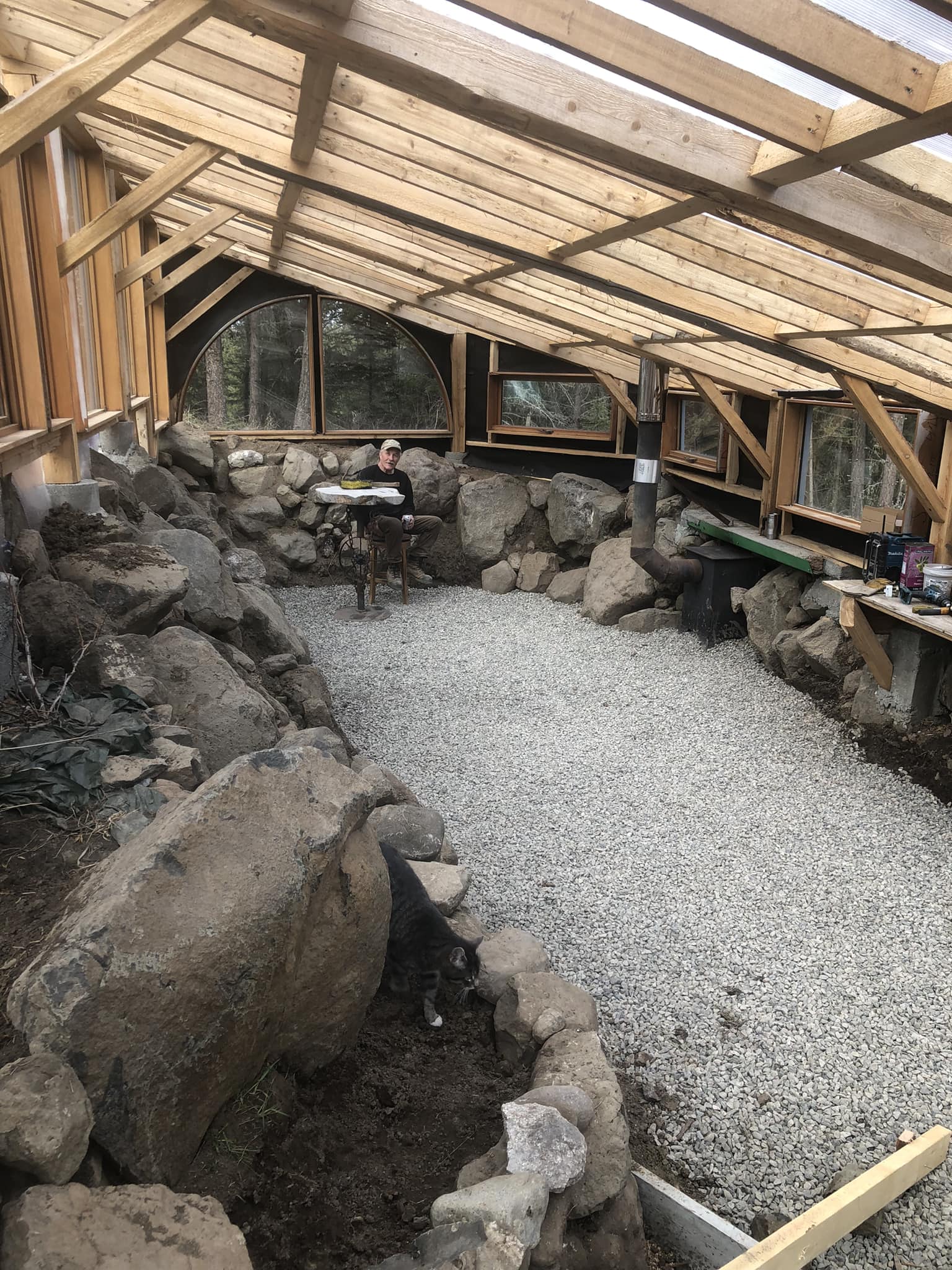 This meme is a photo of a person sitting in a cozy, rustic-style greenhouse made of wood and stone, surrounded by large rocks and a small table. The person is sitting at the table, looking directly at the camera, with a peaceful and contented expression. The text "I finally have my own home" is written above the image, implying that the person has finally found a place to call home, and is feeling happy and content.